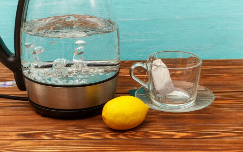 Vařící skleněná konvice, vedle citron a šálek s čajovým sáčkem