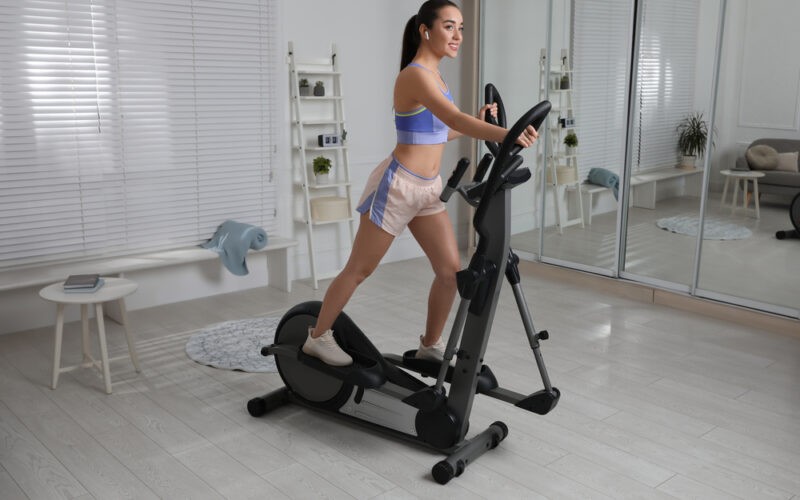 Žena cvičící na eliptickém trenažéru ve středu místnosti před zrcadlem